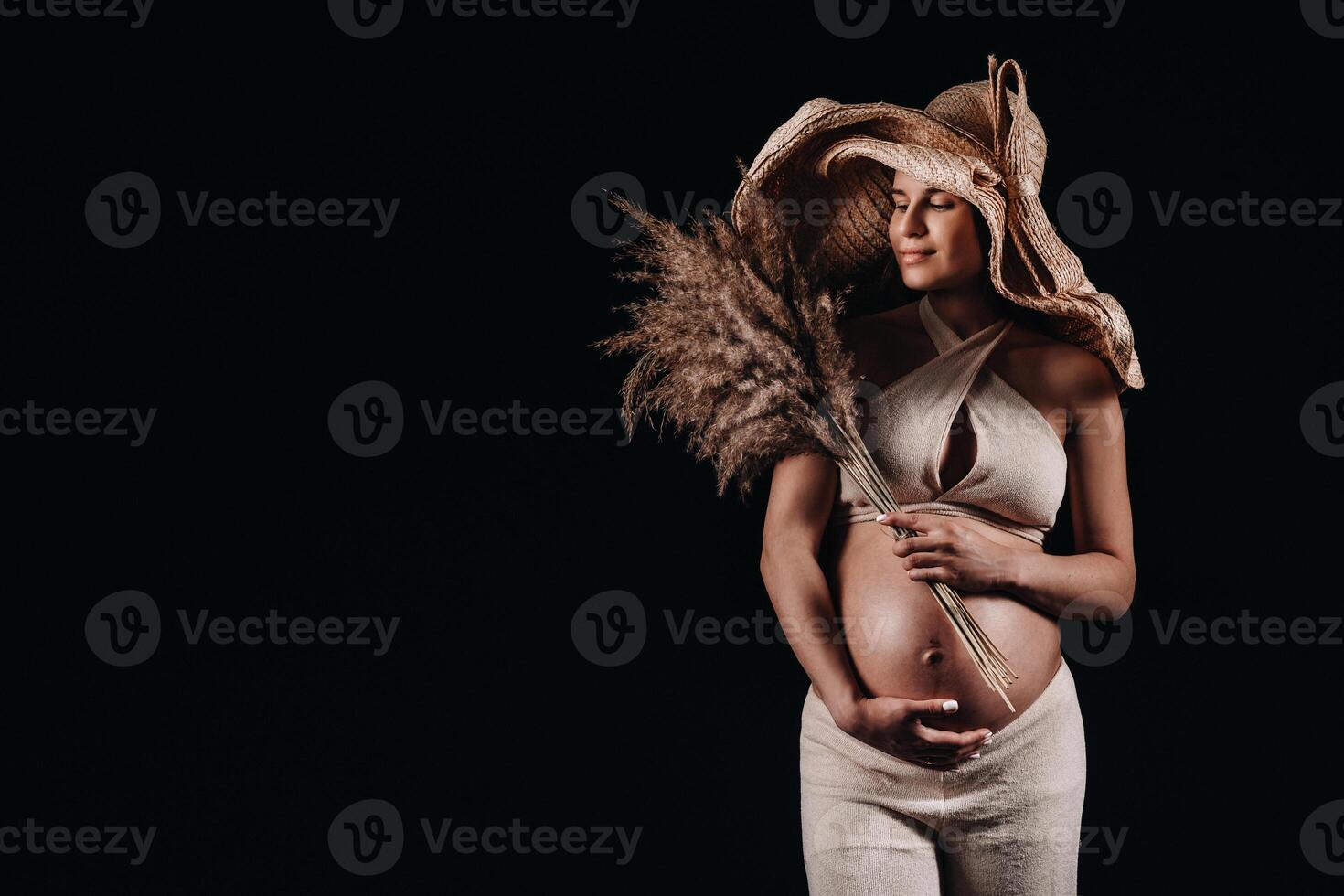 pregnant woman in a straw hat with beige clothes with a bouquet in her hands in the studio on a black background photo