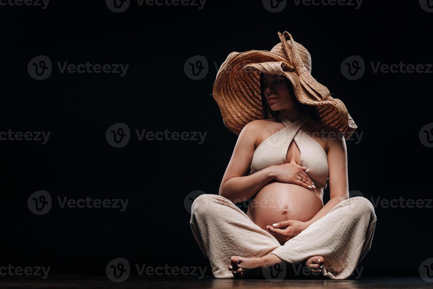 a pregnant woman in a straw hat sits on the floor in beige clothes in a studio on a black background photo
