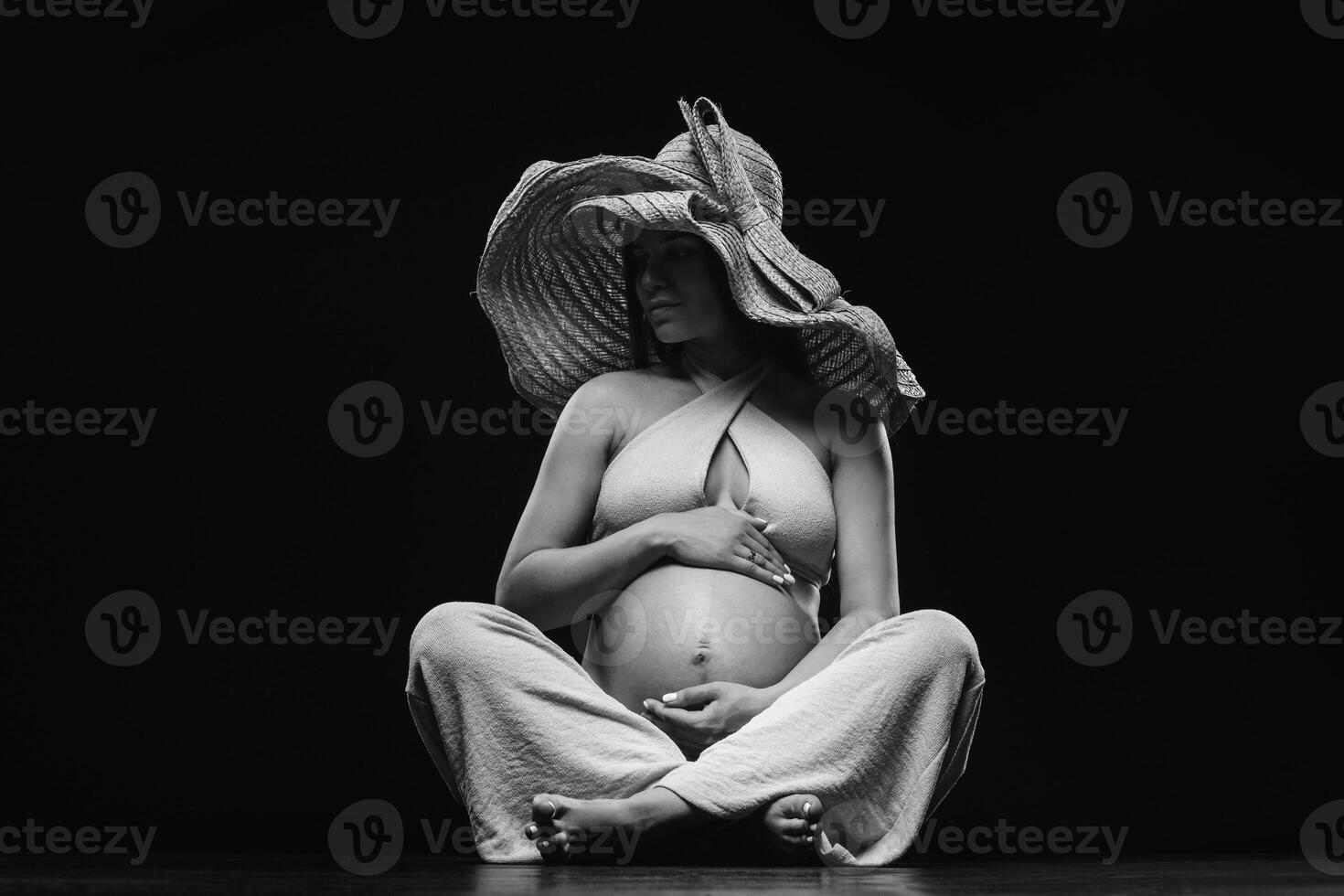 a pregnant woman in a straw hat sits on the floor in beige clothes in a studio on a black background photo