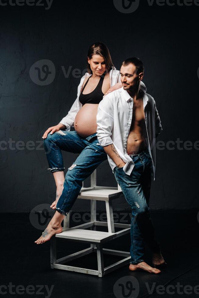 a pregnant woman and a man in a white shirt and jeans in a studio on a black background photo
