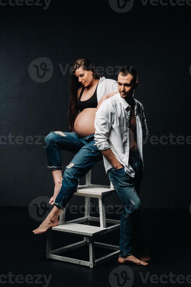 a pregnant woman and a man in a white shirt and jeans in a studio on a black background photo