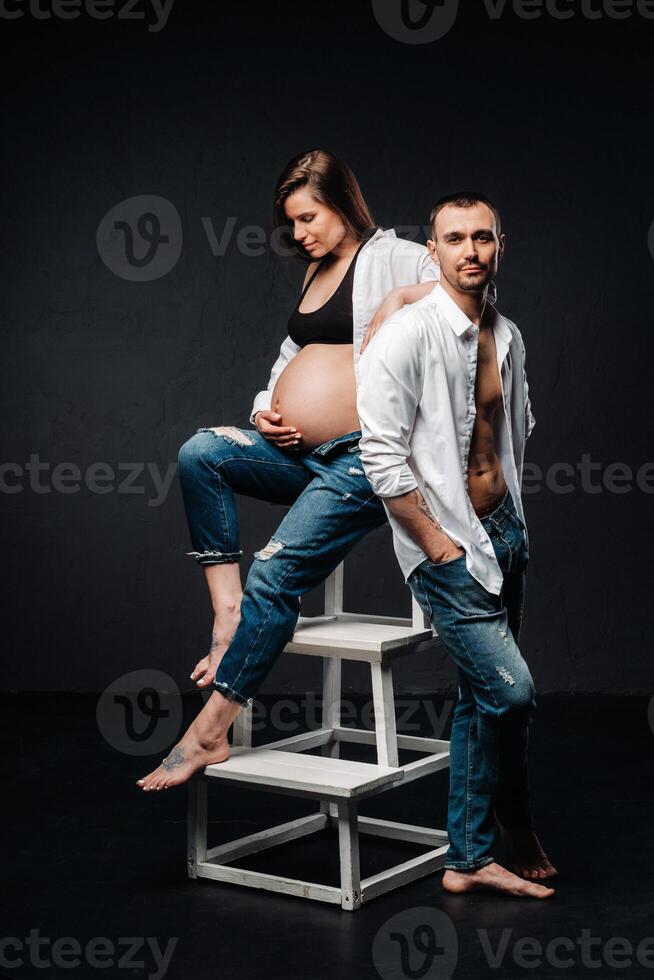 a pregnant woman and a man in a white shirt and jeans in a studio on a black background photo
