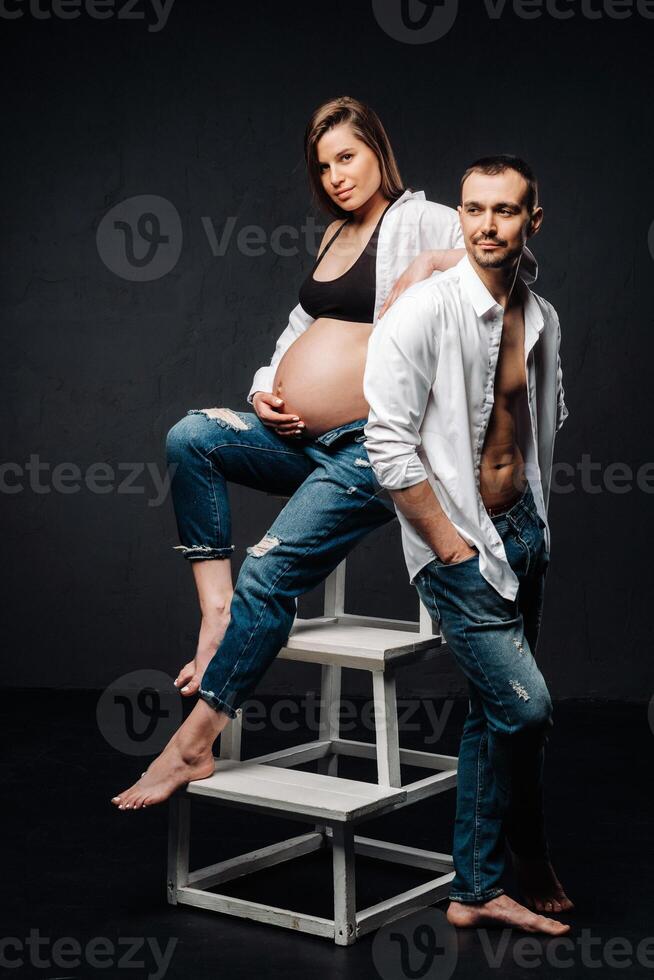 a pregnant woman and a man in a white shirt and jeans in a studio on a black background photo
