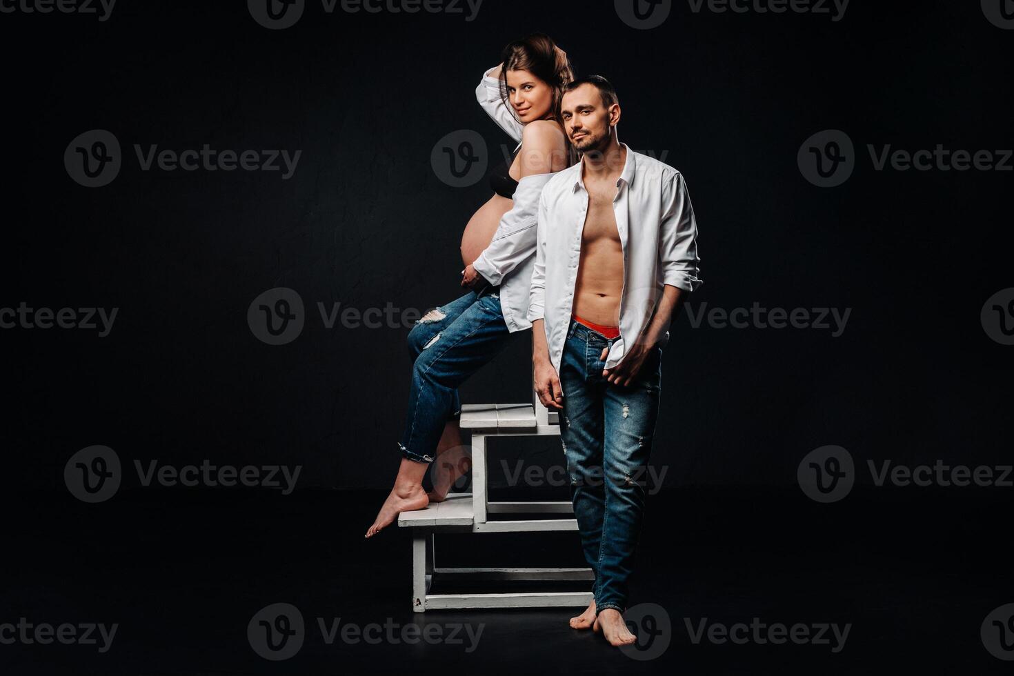 a pregnant woman and a man in a white shirt and jeans in a studio on a black background photo