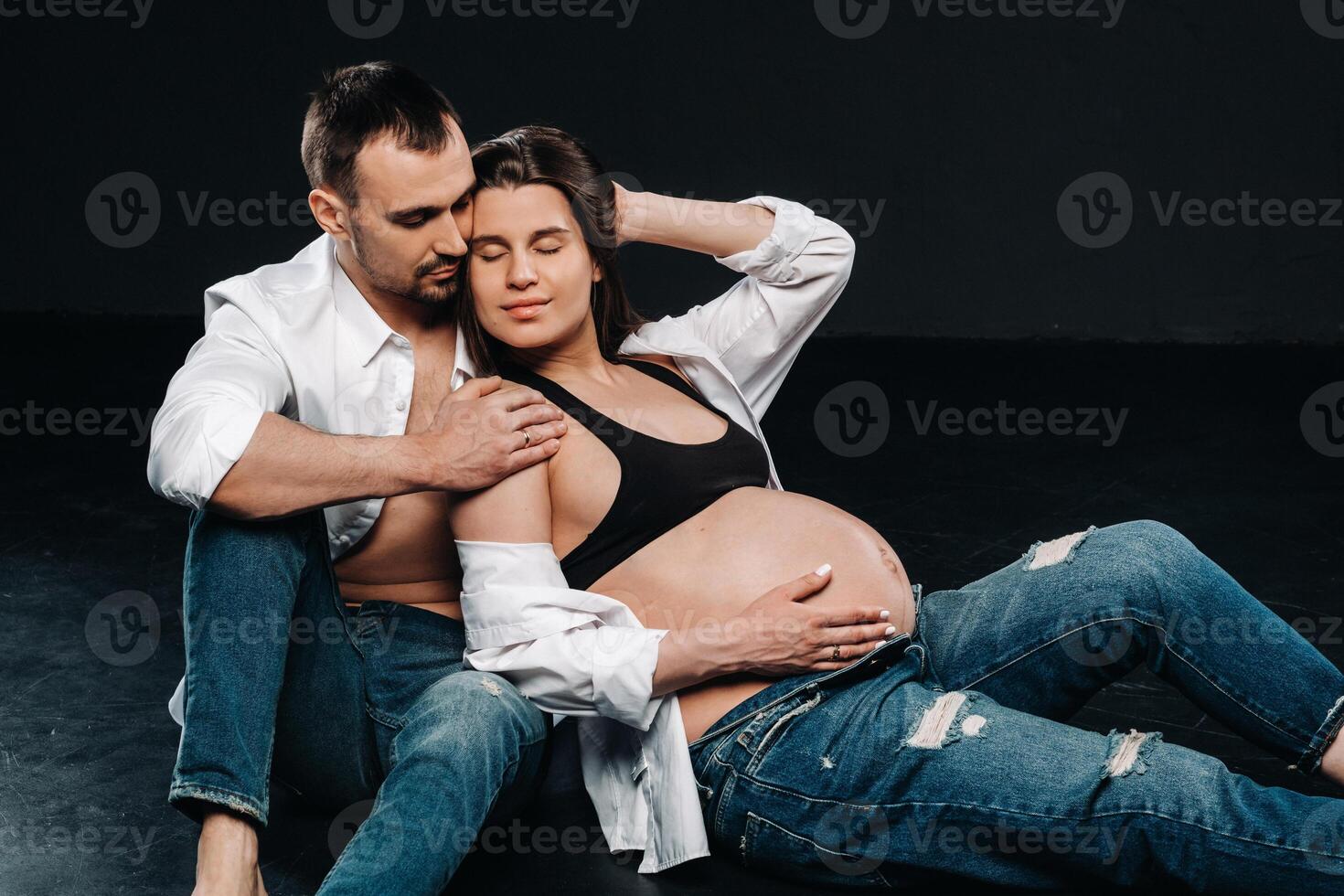 a pregnant woman and a man in a white shirt and jeans in a studio on a black background photo