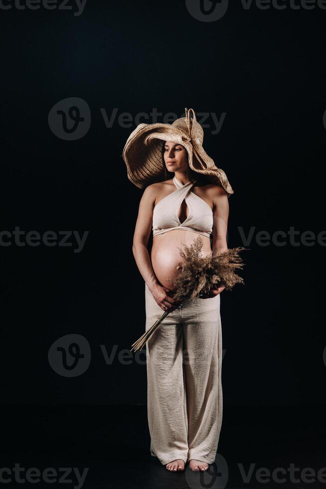 pregnant woman in a straw hat with beige clothes with a bouquet in her hands in the studio on a black background photo