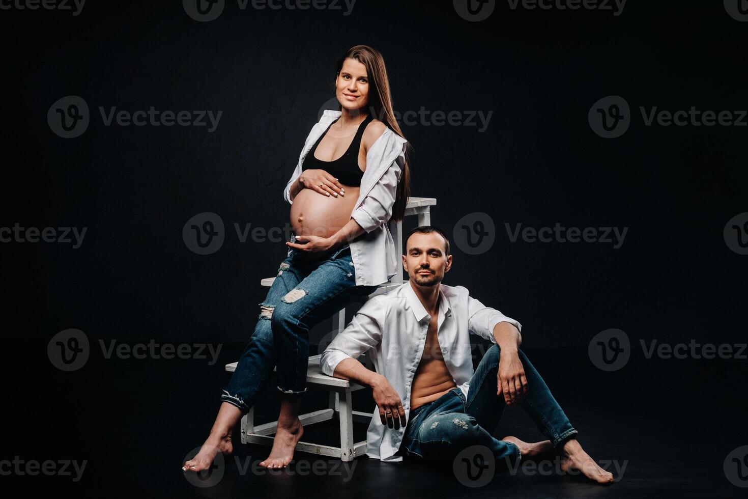 a pregnant woman and a man in a white shirt and jeans in a studio on a black background photo
