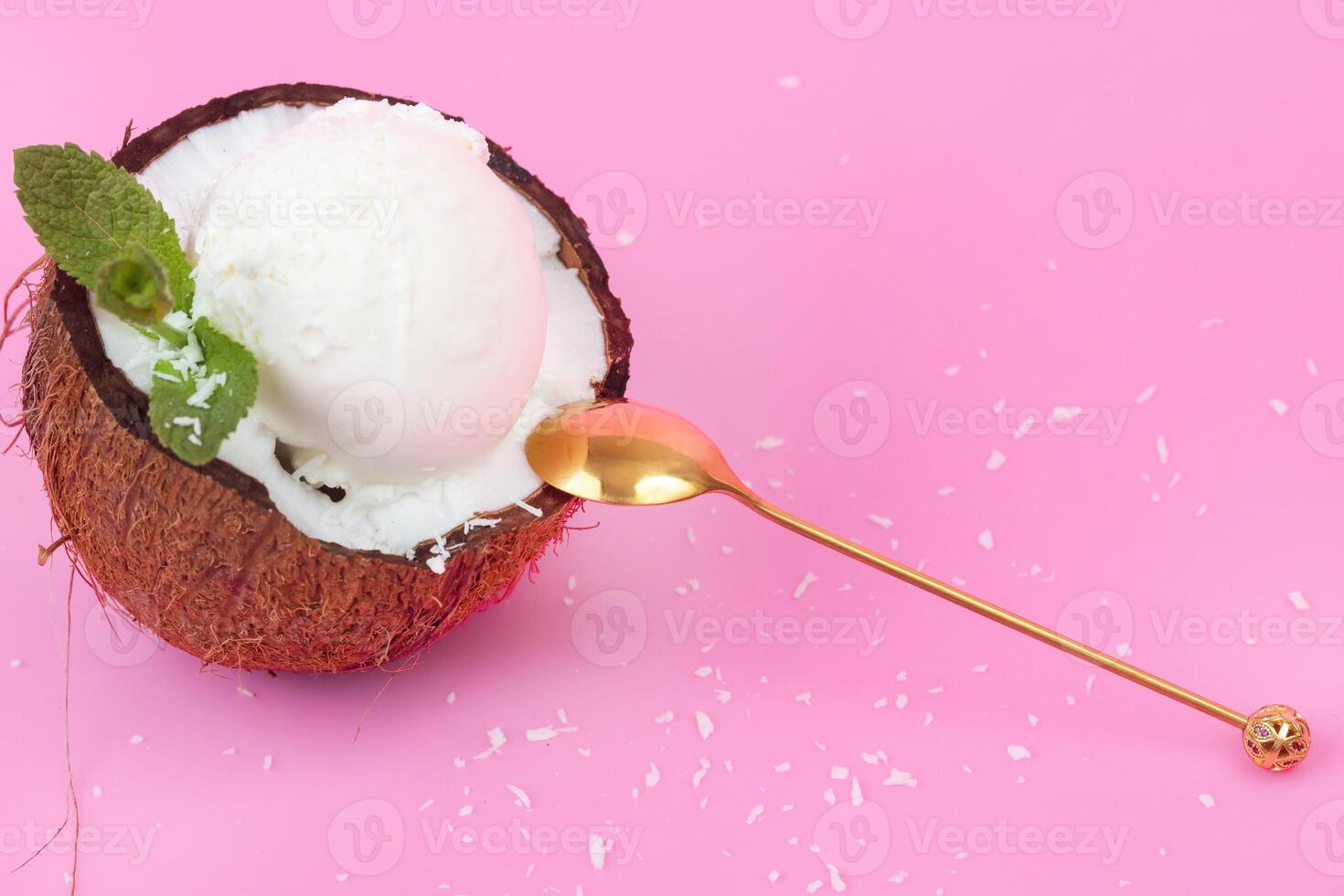 Vanilla ice cream balls in a fresh coconut half decorated with mint leaves with a spoon on a pink background photo