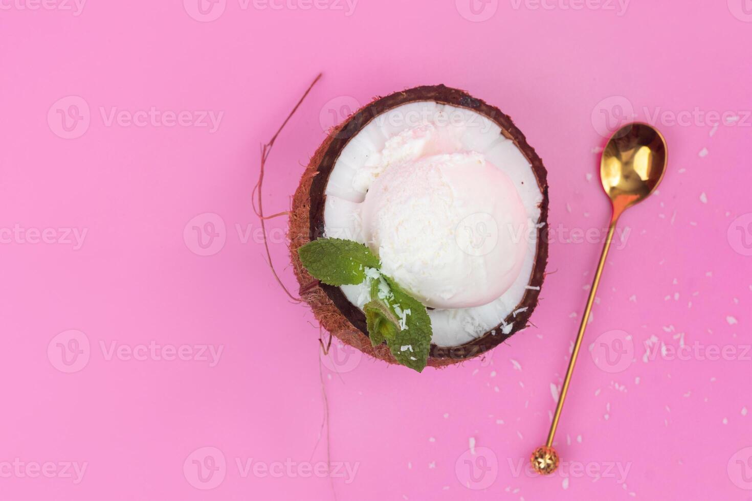 Vanilla ice cream balls in a fresh coconut half decorated with mint leaves with a spoon on a pink background photo