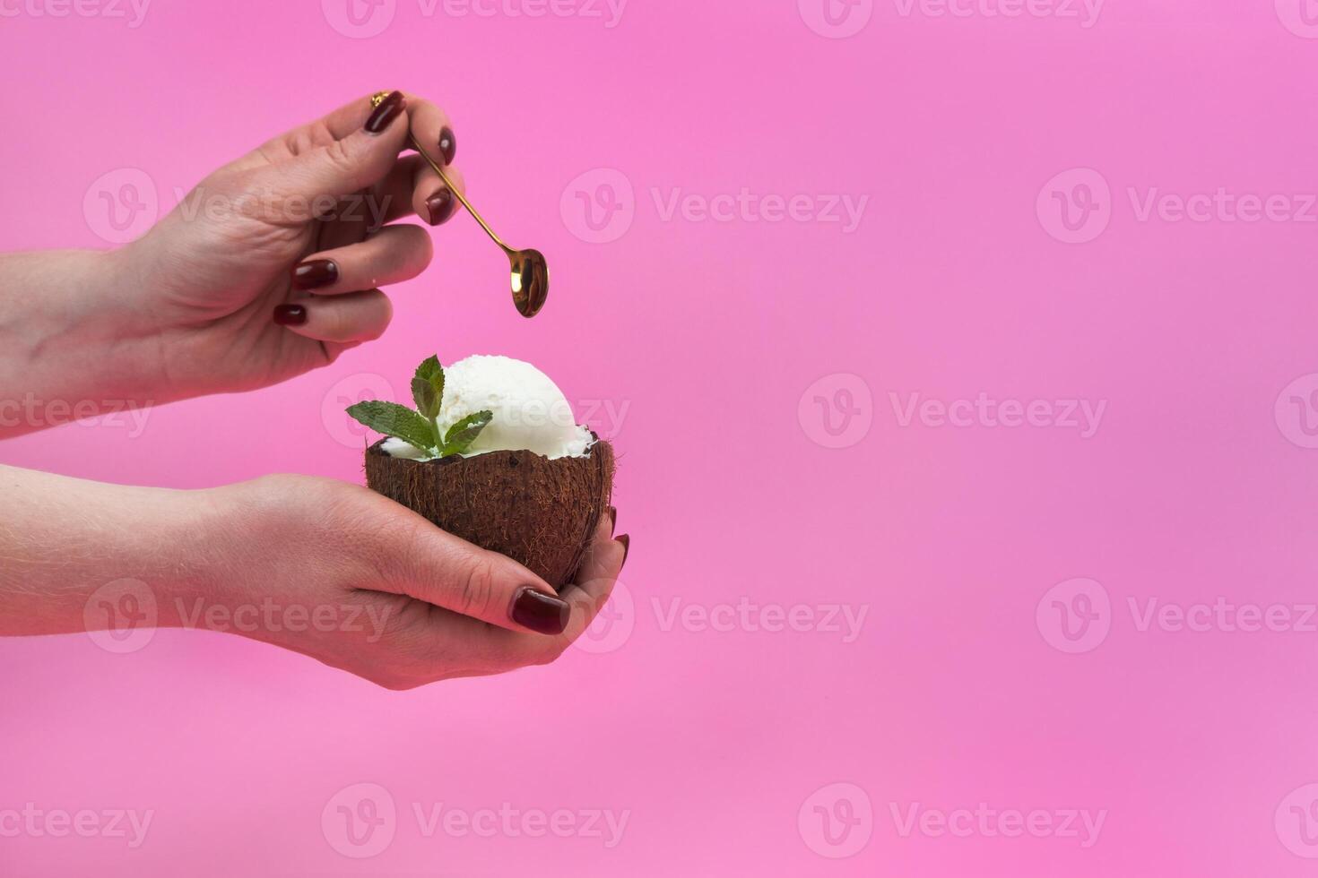 Vanilla ice cream balls in a fresh coconut half, decorated with mint leaves in the hand with a spoon on a pink background photo
