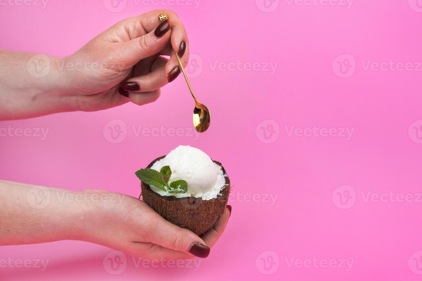 Vanilla ice cream balls in a fresh coconut half, decorated with mint leaves in the hand with a spoon on a pink background photo