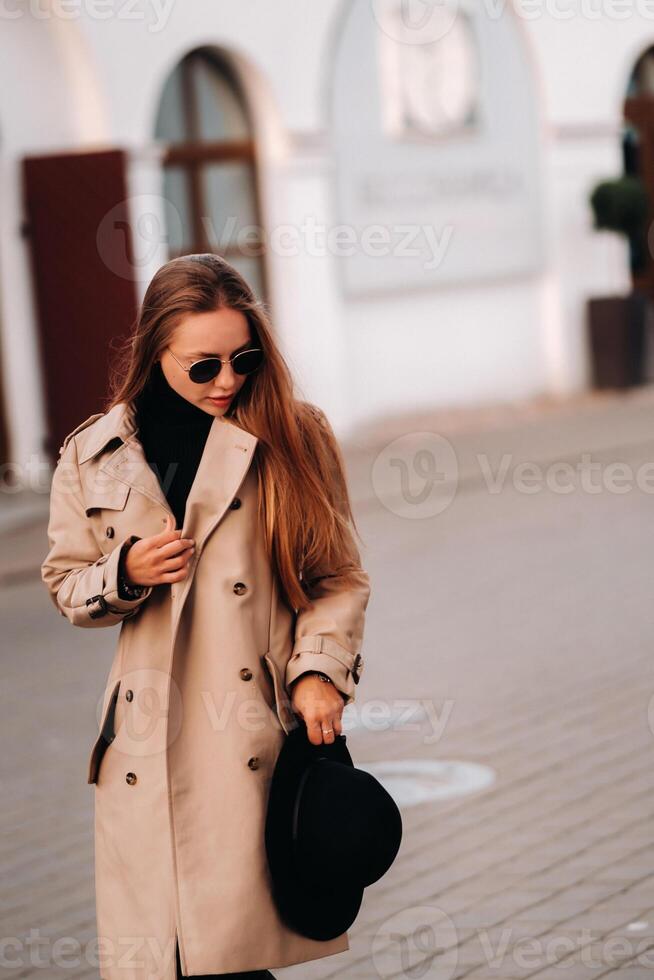 Stylish young woman in a beige coat and a black hat in his hands and glasses on a city street. Women's street fashion. Autumn clothing.Urban style. photo