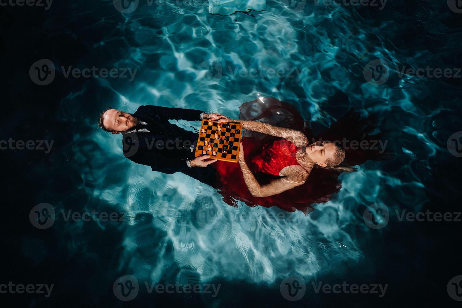 a man in a suit and a girl in a red dress are lying on the water with chess in the pool photo