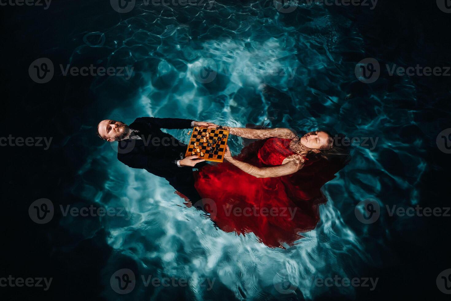 a man in a suit and a girl in a red dress are lying on the water with chess in the pool photo