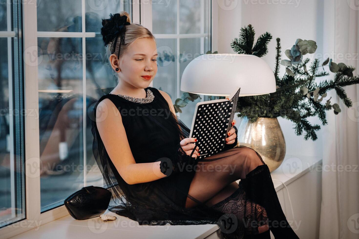 A stylish girl in a black dress sits on the windowsill by the window with a book in her hands photo