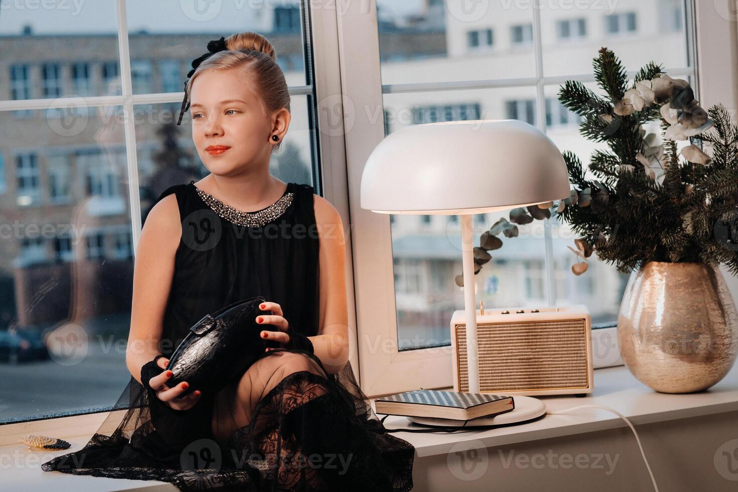 A stylish girl in a black dress sits on the windowsill by the window with a purse in her hands photo