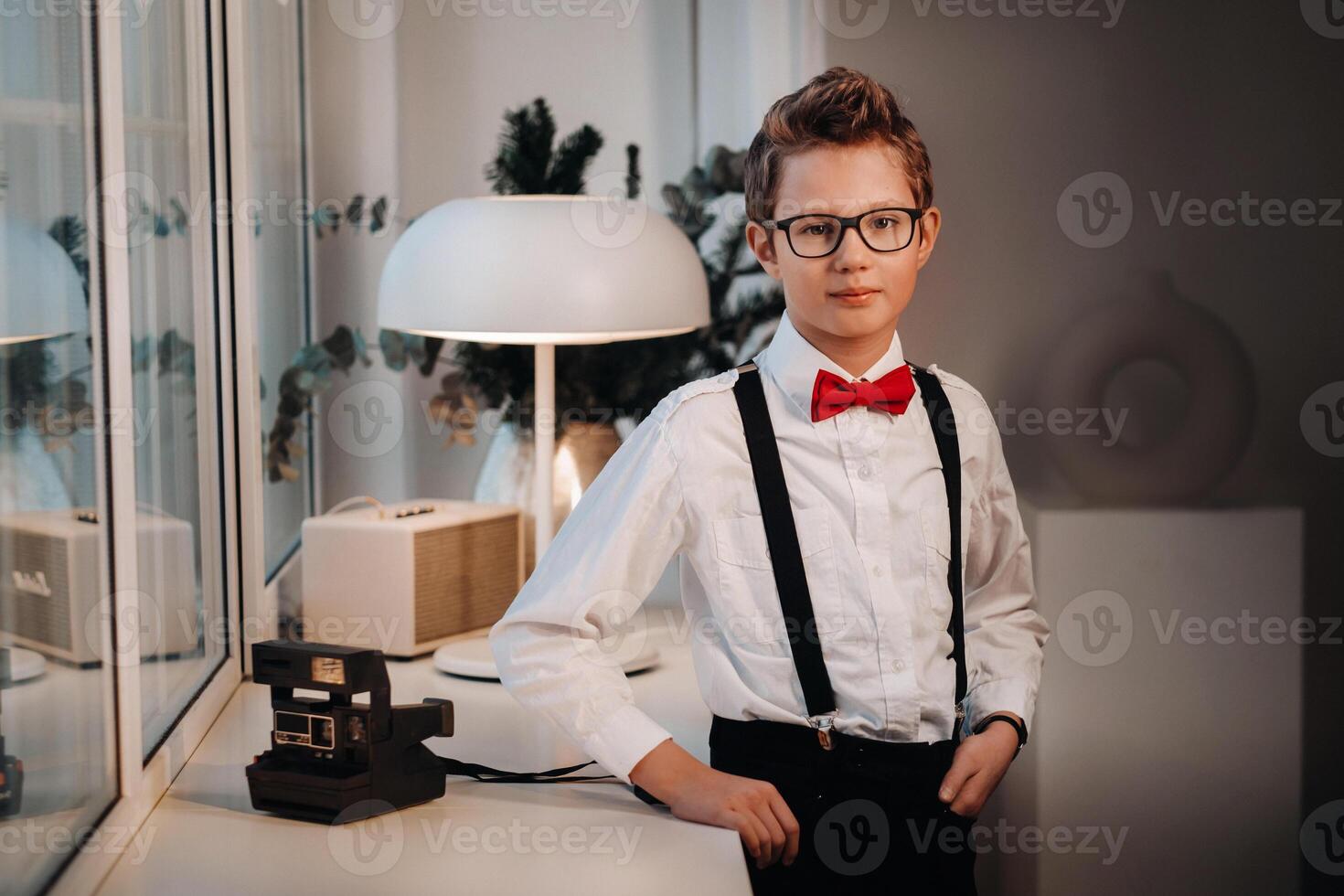 A boy in a shirt with a red bow tie and glasses stands at the window. photo