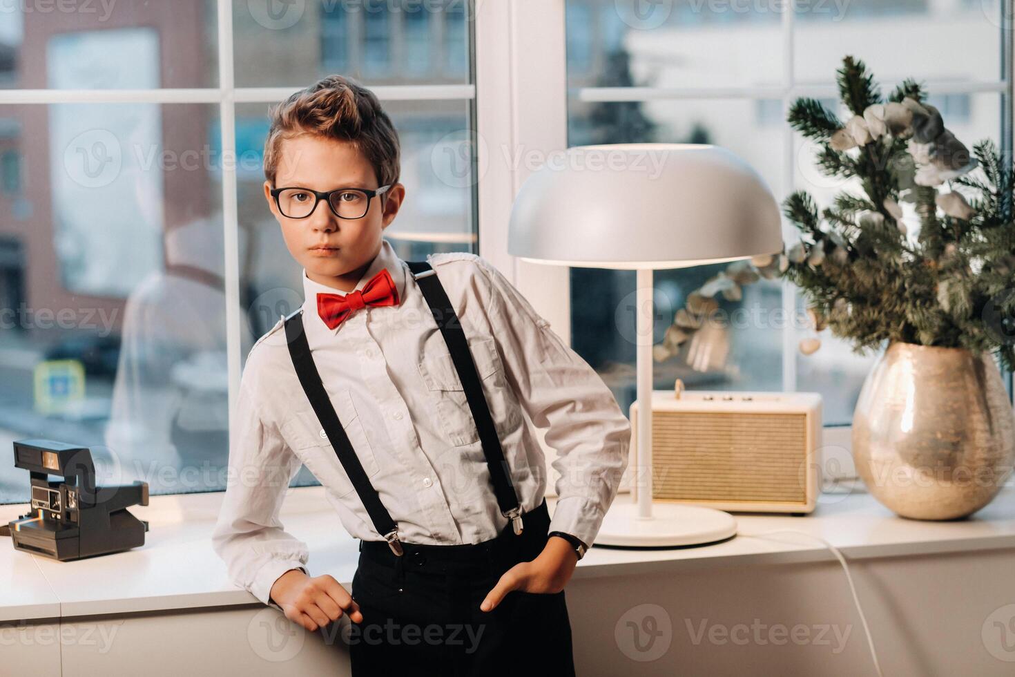 A boy in a shirt with a red bow tie and glasses stands at the window. photo