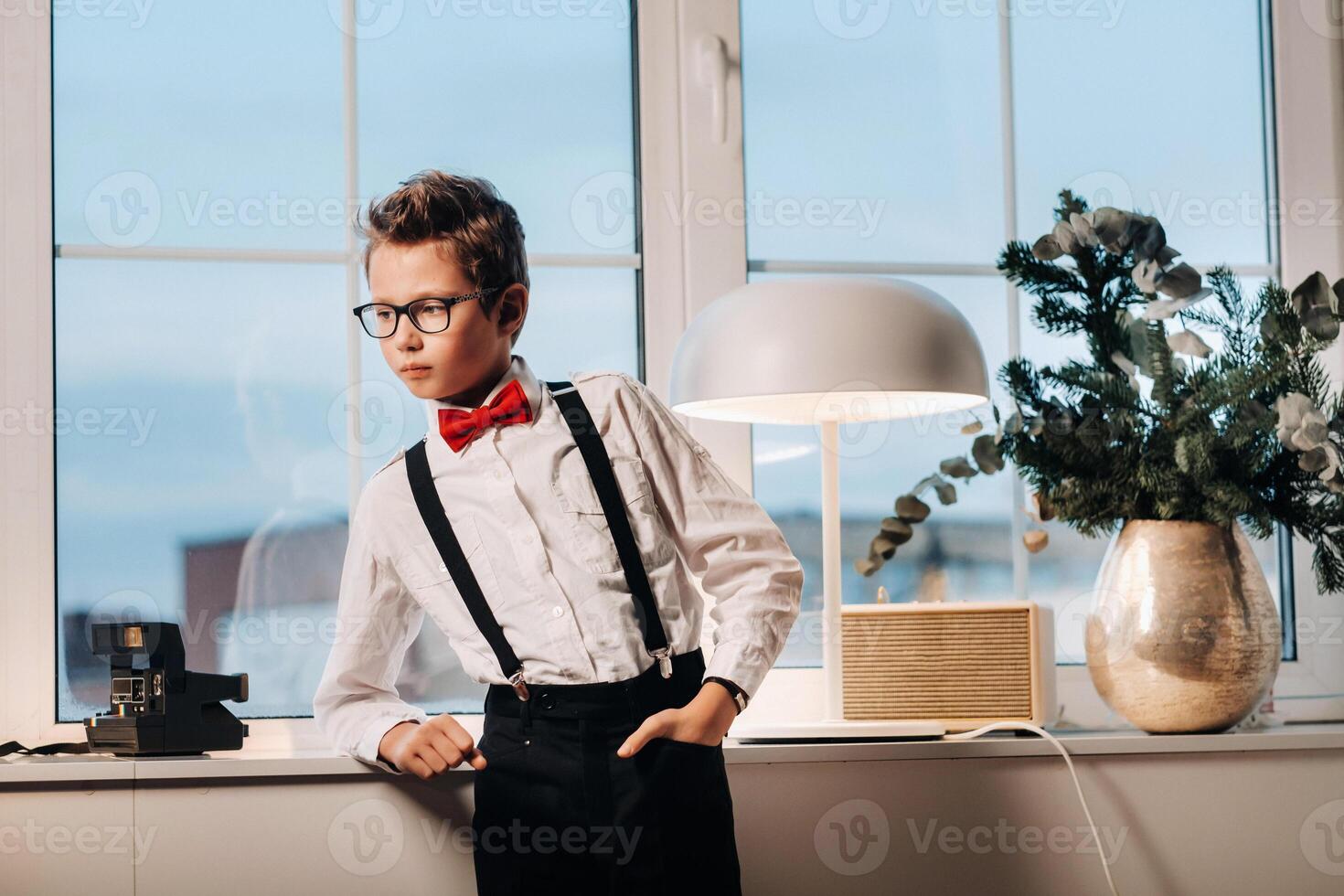 A boy in a shirt with a red bow tie and glasses stands at the window. photo