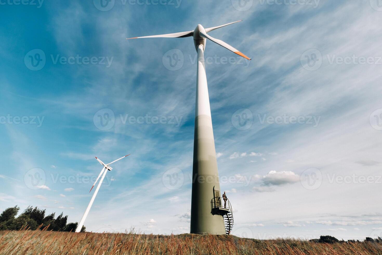 A man in a business suit with a green Golf shirt stands next to a windmill against the background of the field and the blue sky.Businessman near the windmills.Modern concept of the future. photo