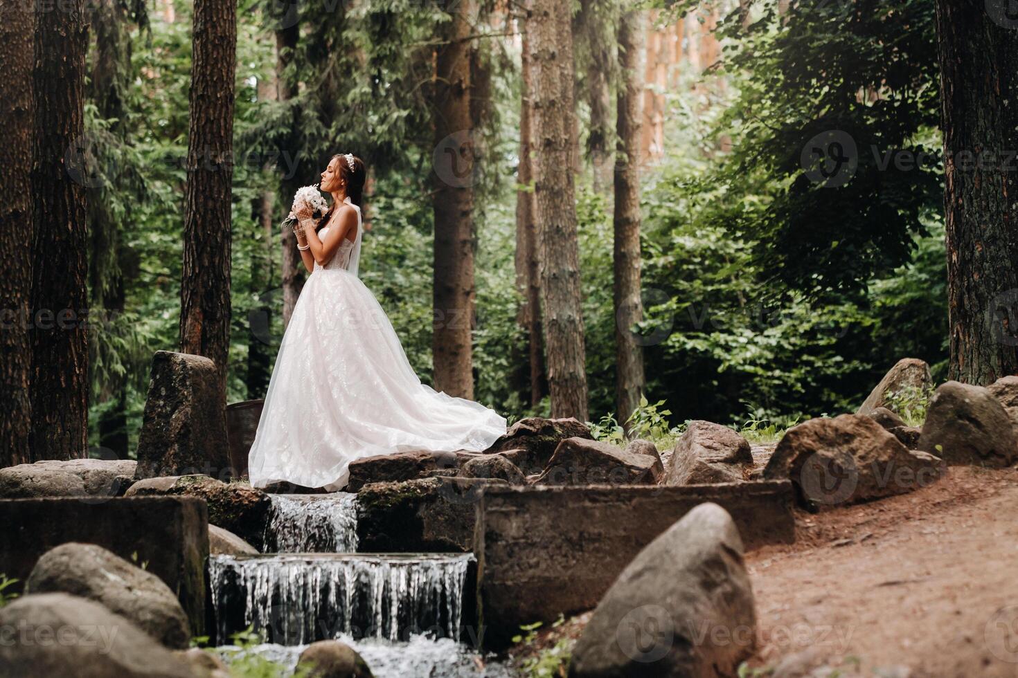An elegant bride in a white dress and gloves holding a bouquet stands by a stream in the forest, enjoying nature.A model in a wedding dress and gloves in a nature Park.Belarus photo