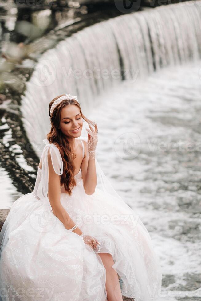 An elegant bride in a white dress, gloves and bare feet is sitting near a waterfall in the Park enjoying nature.A model in a wedding dress and gloves at a nature Park.Belarus photo