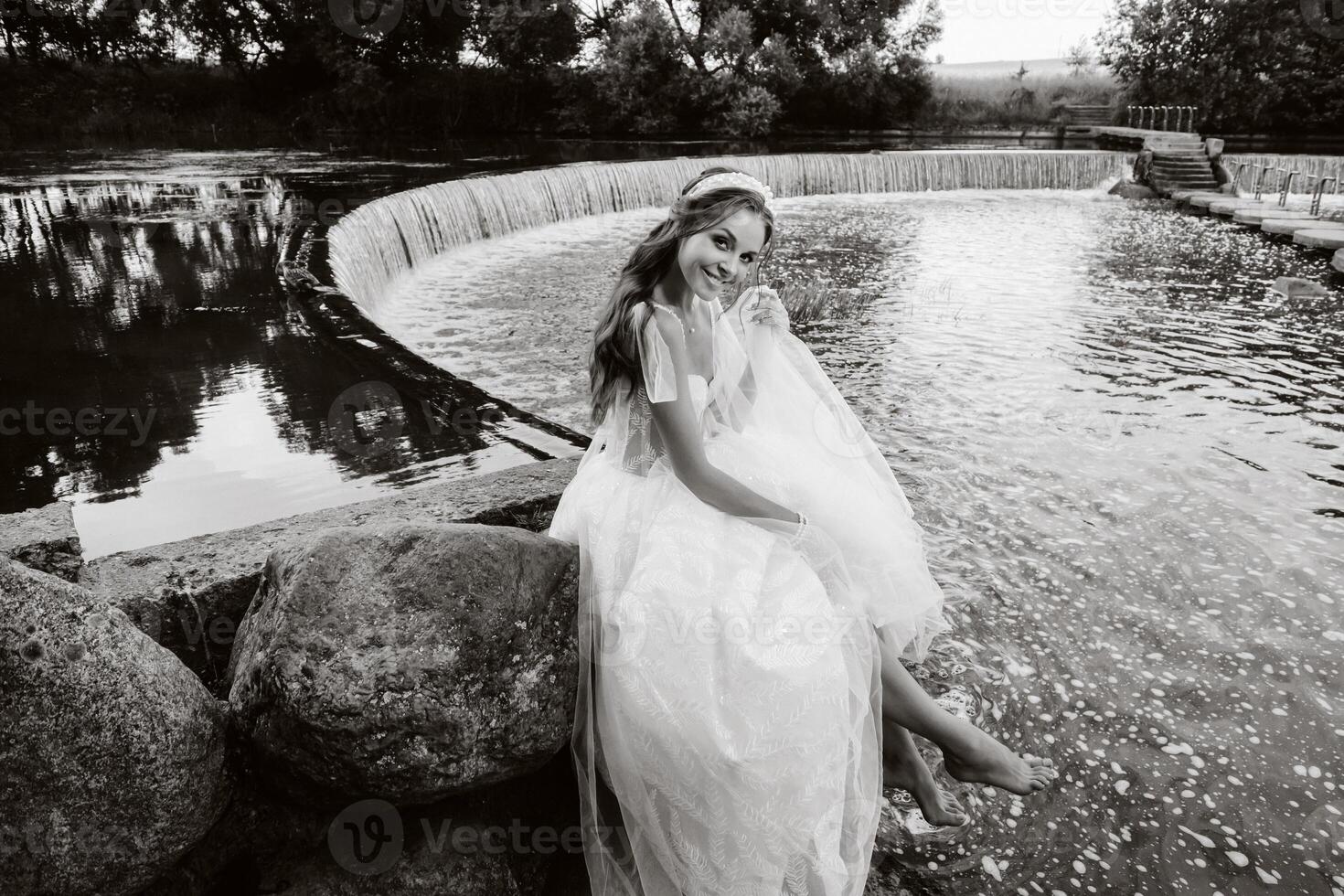 An elegant bride in a white dress, gloves and bare feet is sitting near a waterfall in the Park enjoying nature.A model in a wedding dress and gloves at a nature Park.Belarus photo