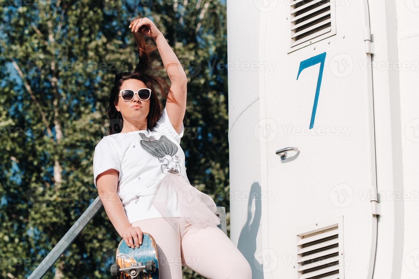 A girl in white clothes and glasses with a skate in her hands is photographed near large wind turbines in a field with trees.Modern woman with a riding Board in a field with windmills photo