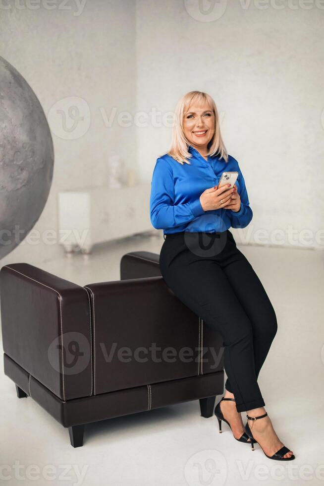 A fashionable woman in a blue shirt and black trousers poses in the interior with a smartphone in her hands photo