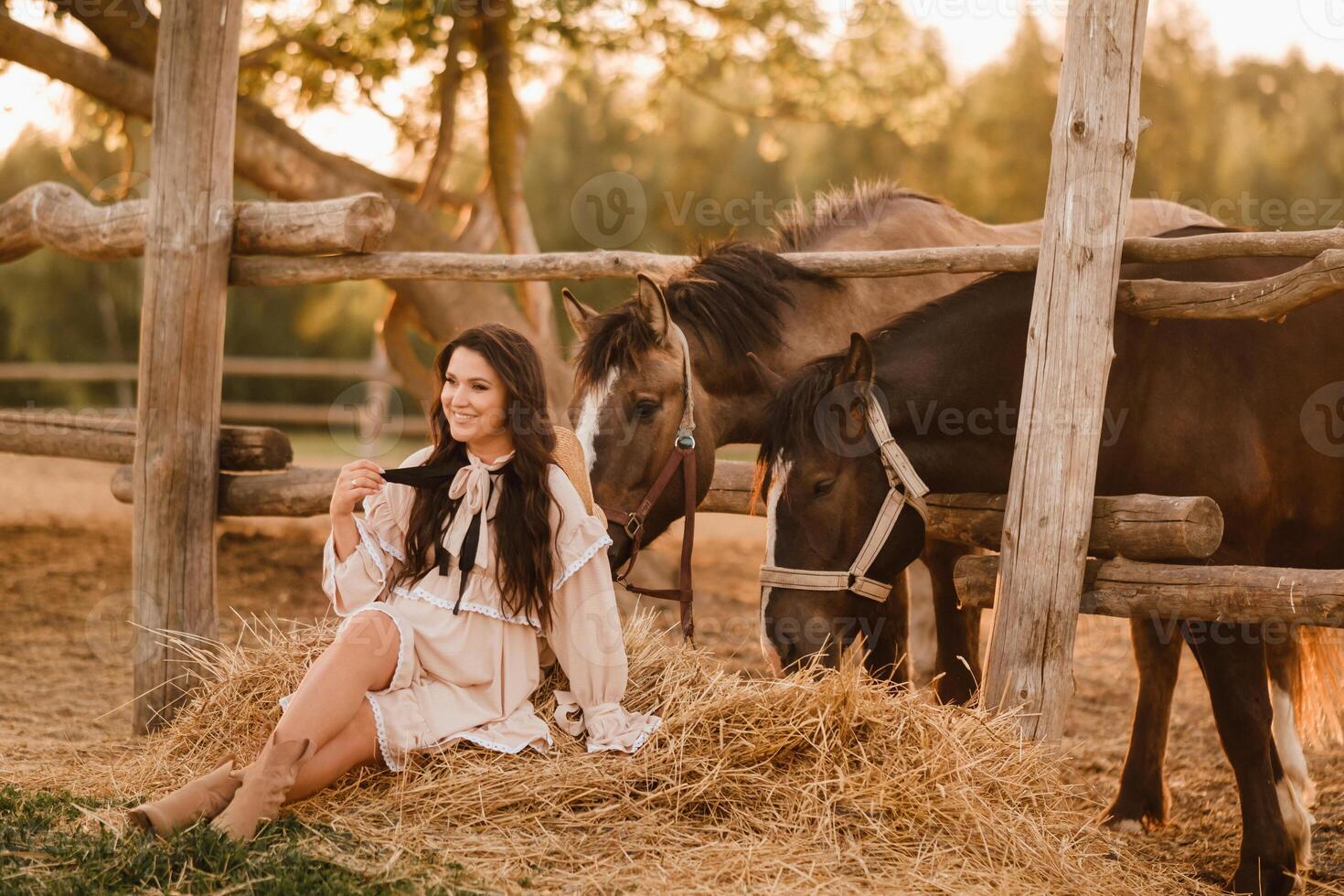 a pregnant woman in a dress in the countryside is sitting on the hay near the horses photo