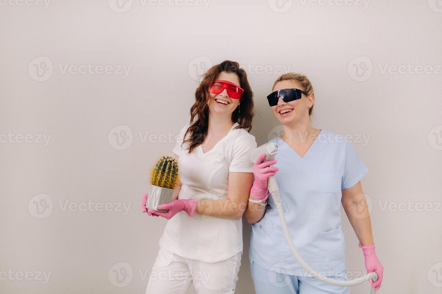 Two cosmetologists in protective glasses are holding a laser depilation device and a cactus in their hands photo