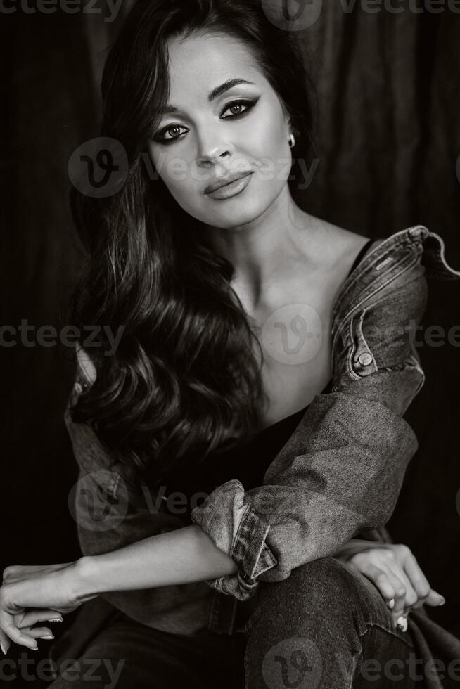 a brunette with long hair poses in the studio sitting on the floor on a black background.Black and white photo