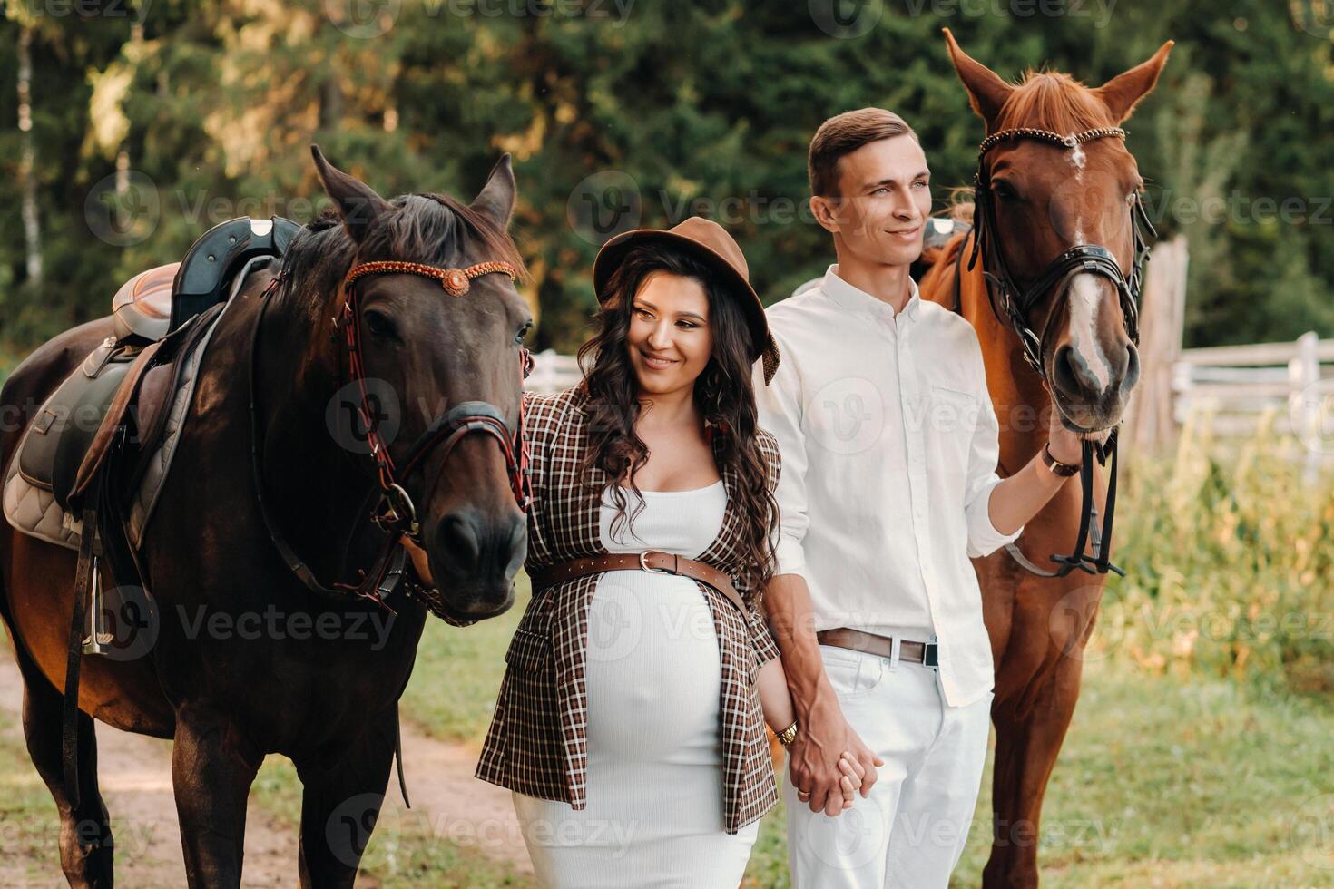 a pregnant girl in a hat and her husband in white clothes stand next to horses in the forest in nature.Stylish pregnant woman with a man with horses.Family. photo