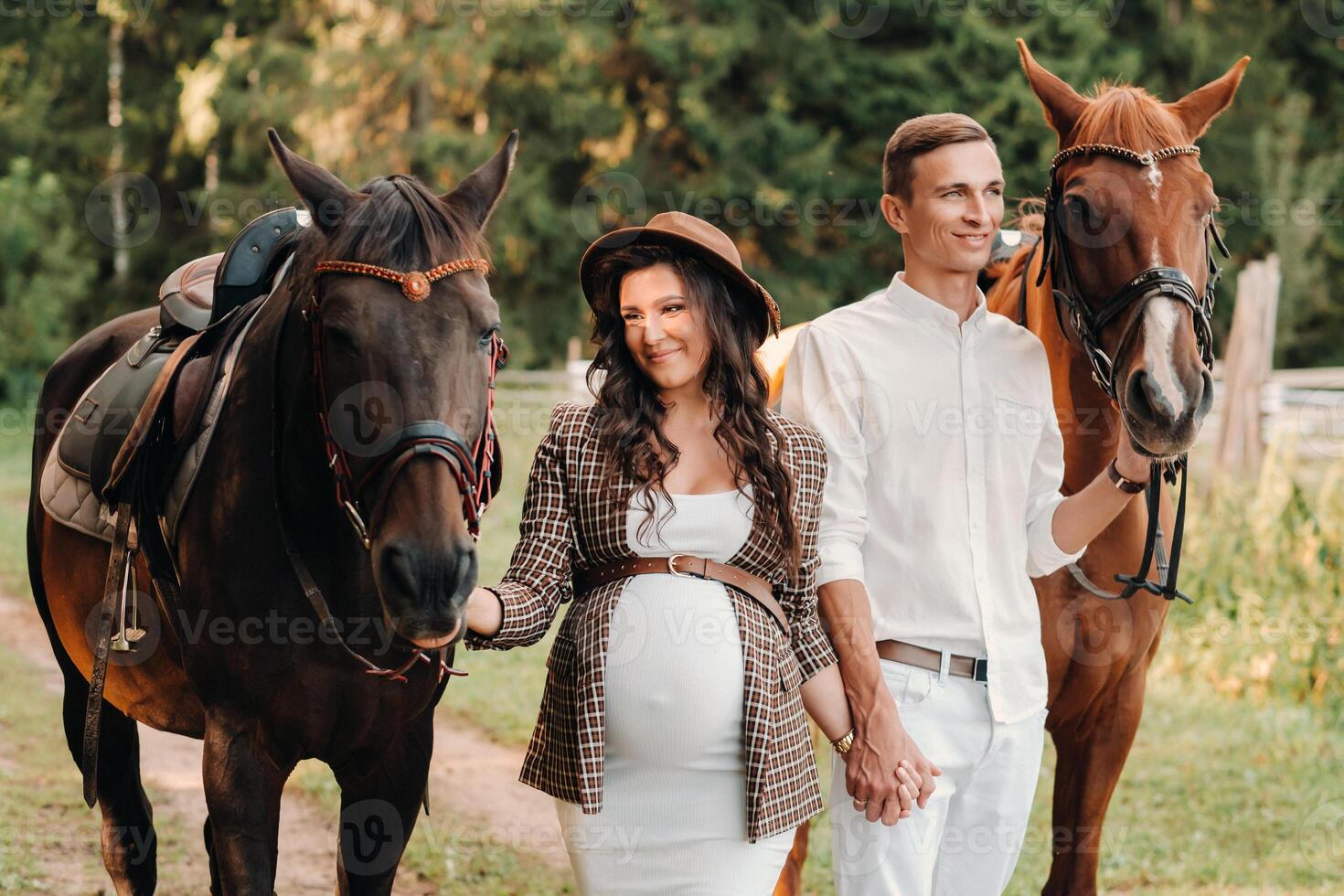 A pregnant woman in a hat with a man in white clothes walking with horses in nature. A family waiting for a child walks in the woods photo