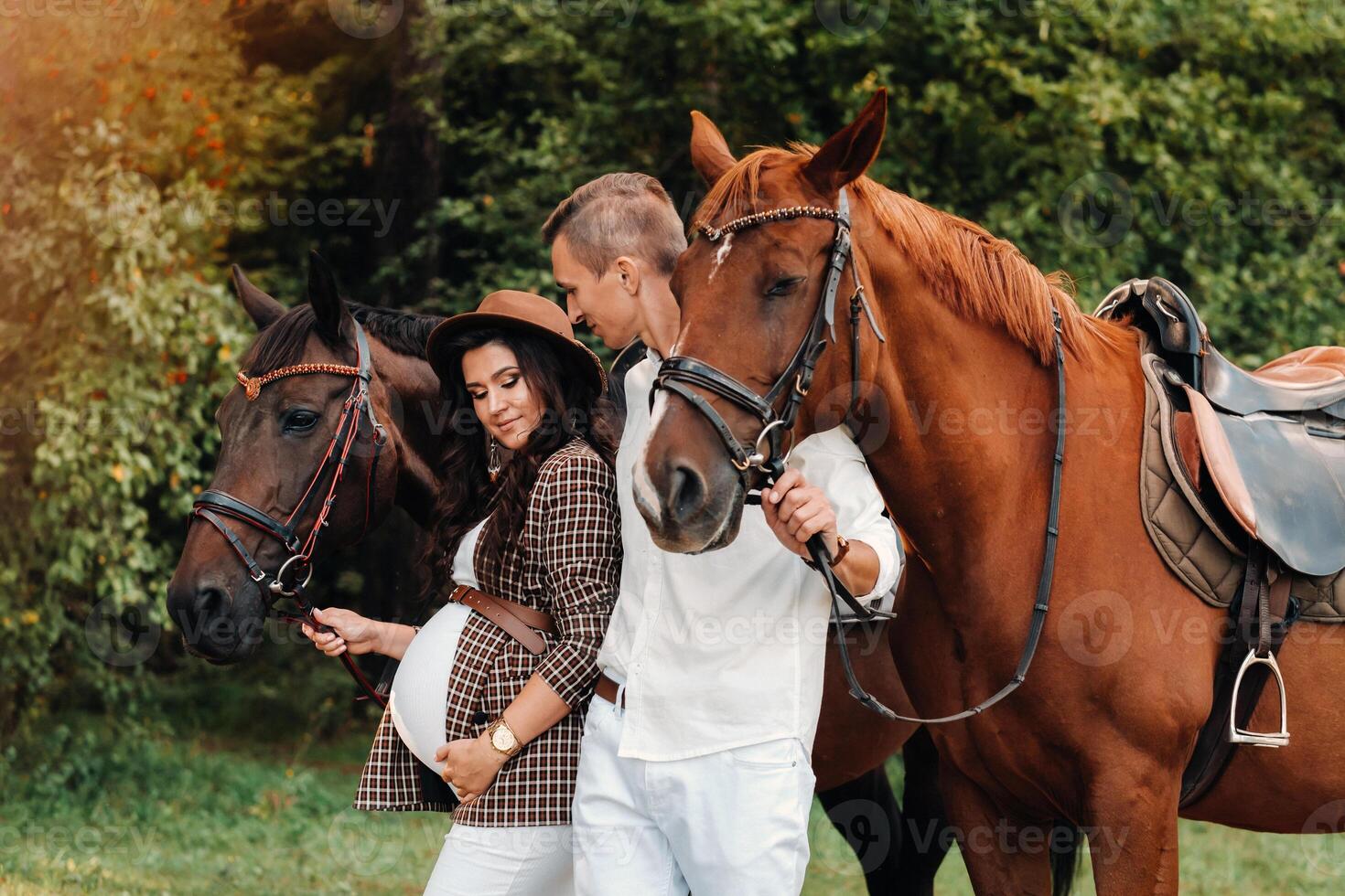 a pregnant girl in a hat and her husband in white clothes stand next to horses in the forest in nature.Stylish pregnant woman with a man with horses.Family. photo