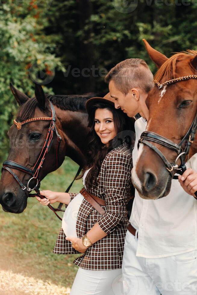a pregnant girl in a hat and her husband in white clothes stand next to horses in the forest in nature.Stylish pregnant woman with a man with horses.Family. photo