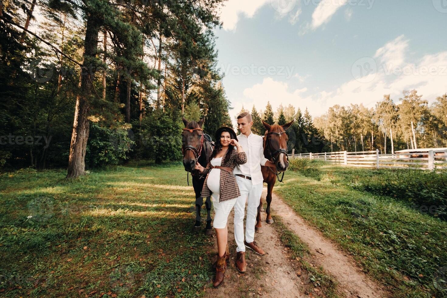 a pregnant girl in a hat and her husband in white clothes stand next to horses in the forest in nature.Stylish pregnant woman with a man with horses.Family. photo