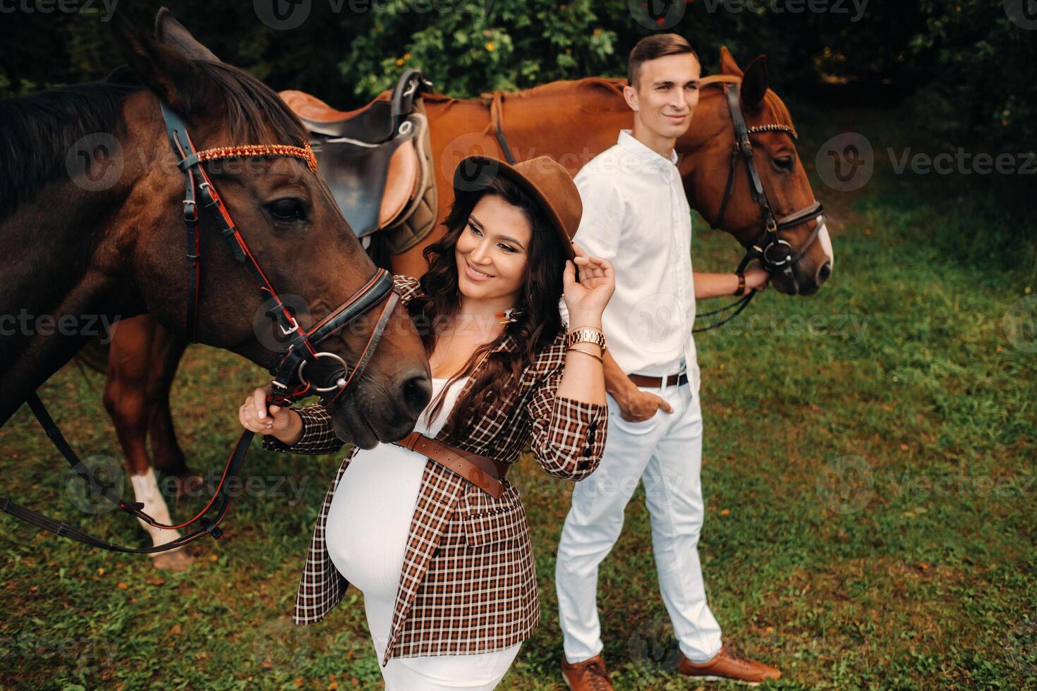 A pregnant woman in a hat with a man in white clothes walking with horses in nature. A family waiting for a child walks in the woods photo