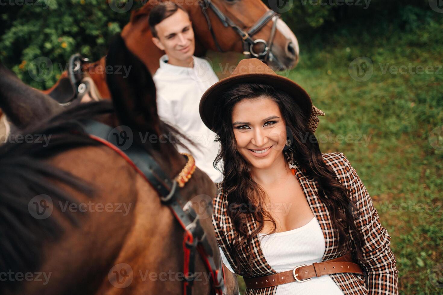 a pregnant girl in a hat and a man in white clothes stand next to horses in the forest in nature.Stylish pregnant woman with her husband with horses.Married couple. photo