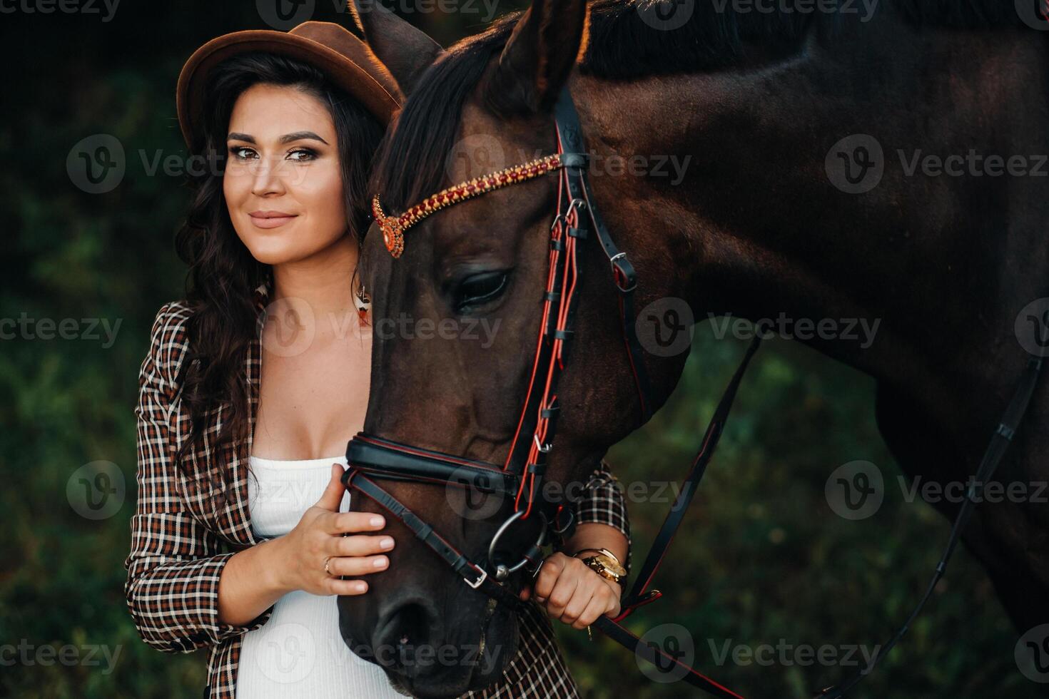 pregnant girl with a big belly in a hat next to horses in the forest in nature.Stylish girl in white clothes and a brown jacket. photo