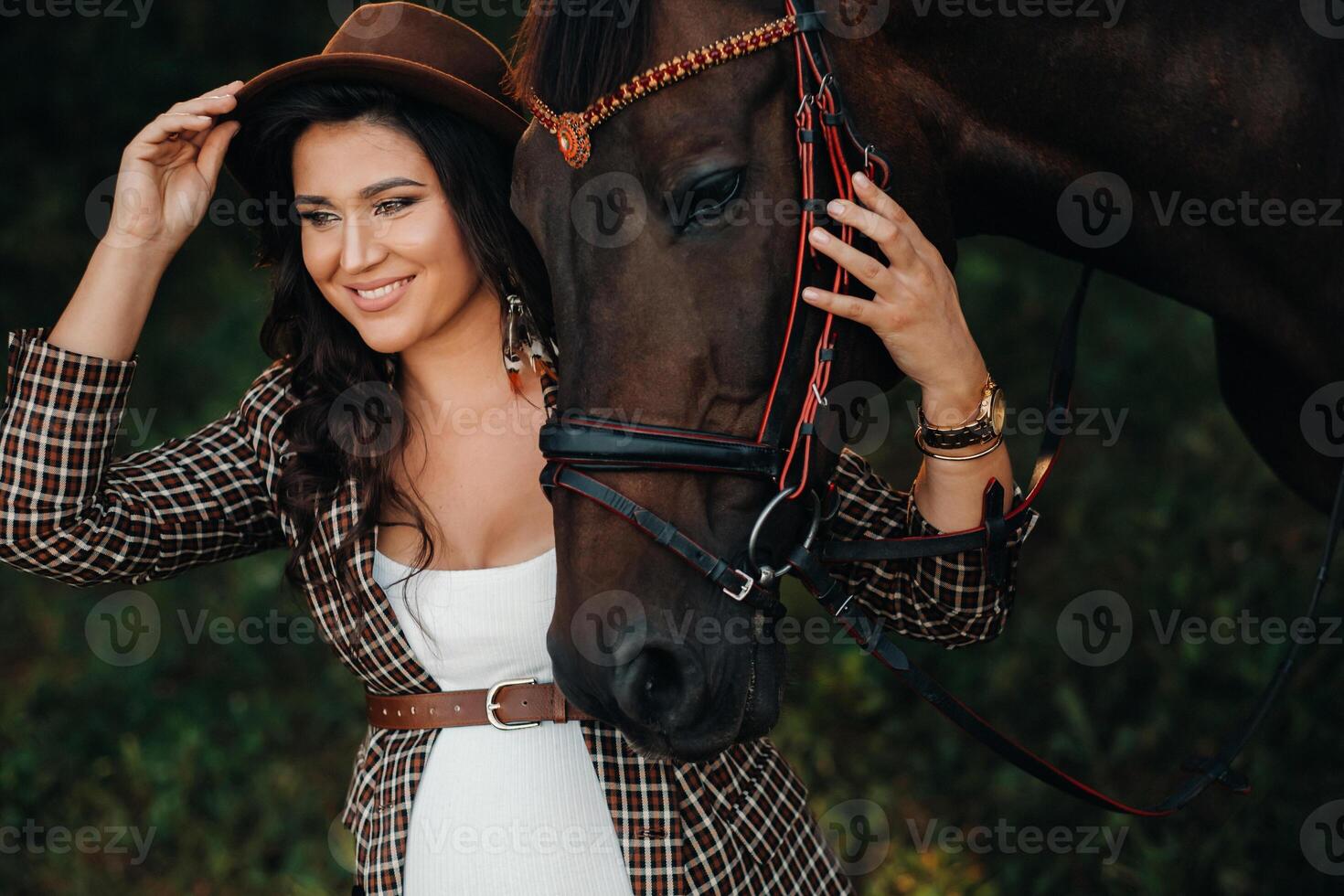 pregnant girl with a big belly in a hat next to horses in the forest in nature.Stylish girl in white clothes and a brown jacket. photo