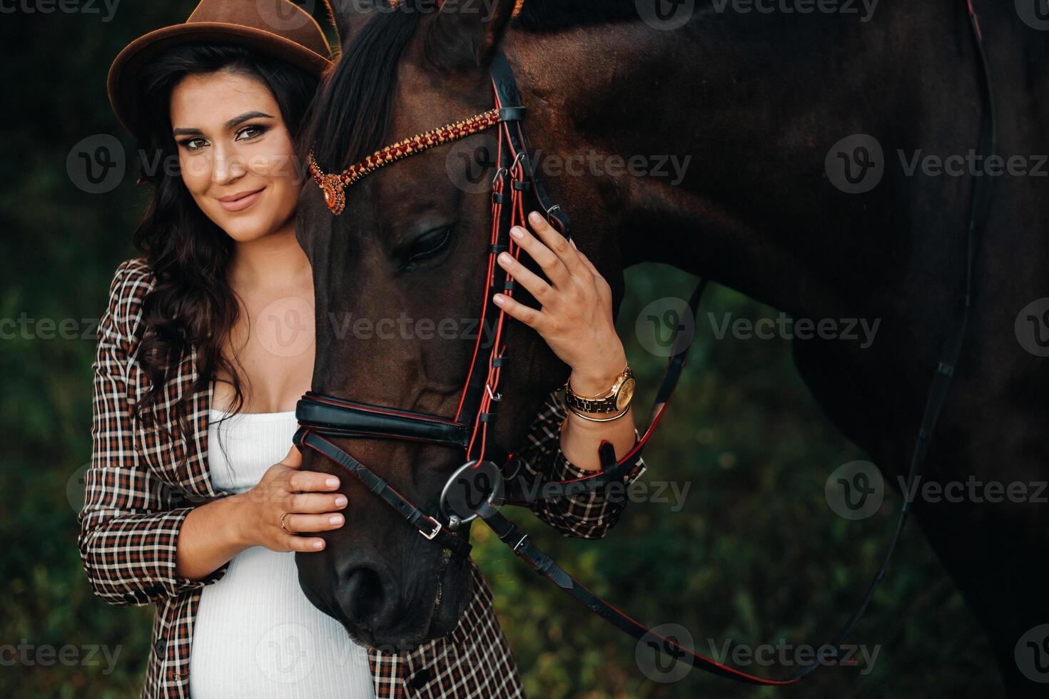pregnant girl with a big belly in a hat next to horses in the forest in nature.Stylish girl in white clothes and a brown jacket. photo