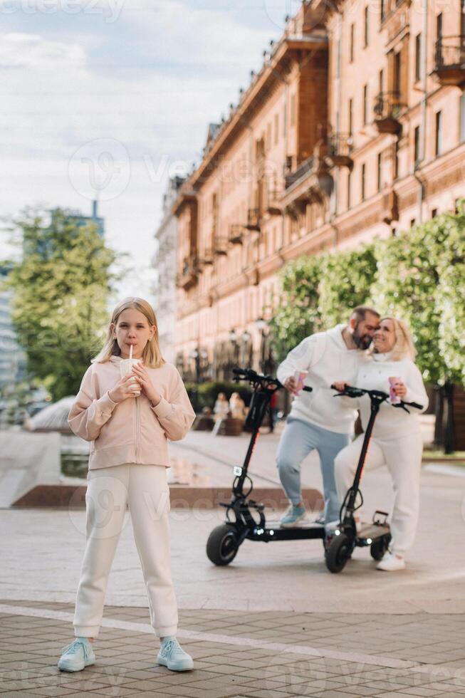 A girl with a drink in her hands is standing in the city and behind her on electric scooters are her parents photo
