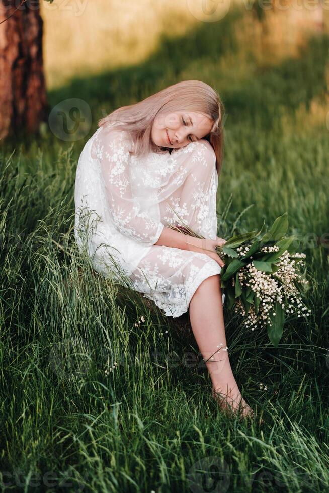 A beautiful nine-year-old blonde with long hair in a long white dress, holding a bouquet of lilies of the valley, sits on a stone in the Park.Summer, sunset. photo