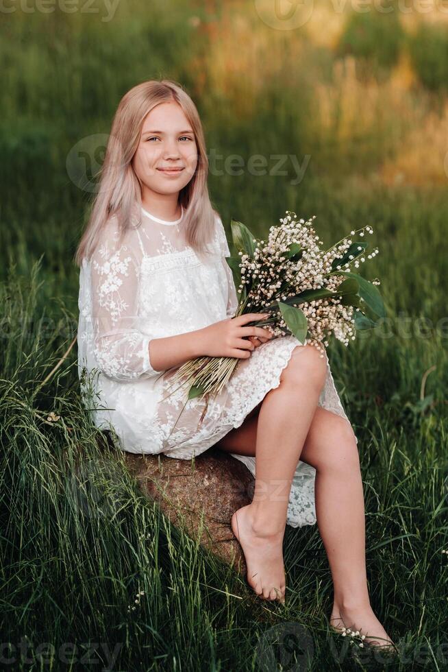 A beautiful nine-year-old blonde girl with long hair in a long white dress, holding a bouquet of lilies of the valley flowers, walking in nature in the Park.Summer, sunset. photo