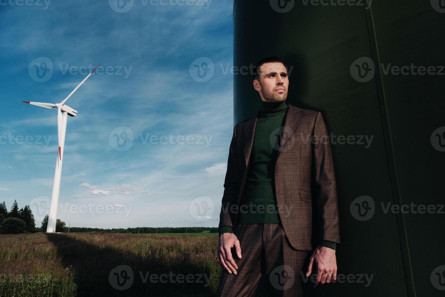 A man in a business suit with a green Golf shirt stands next to a windmill against the background of the field and the blue sky.Businessman near the windmills.Modern concept of the future. photo