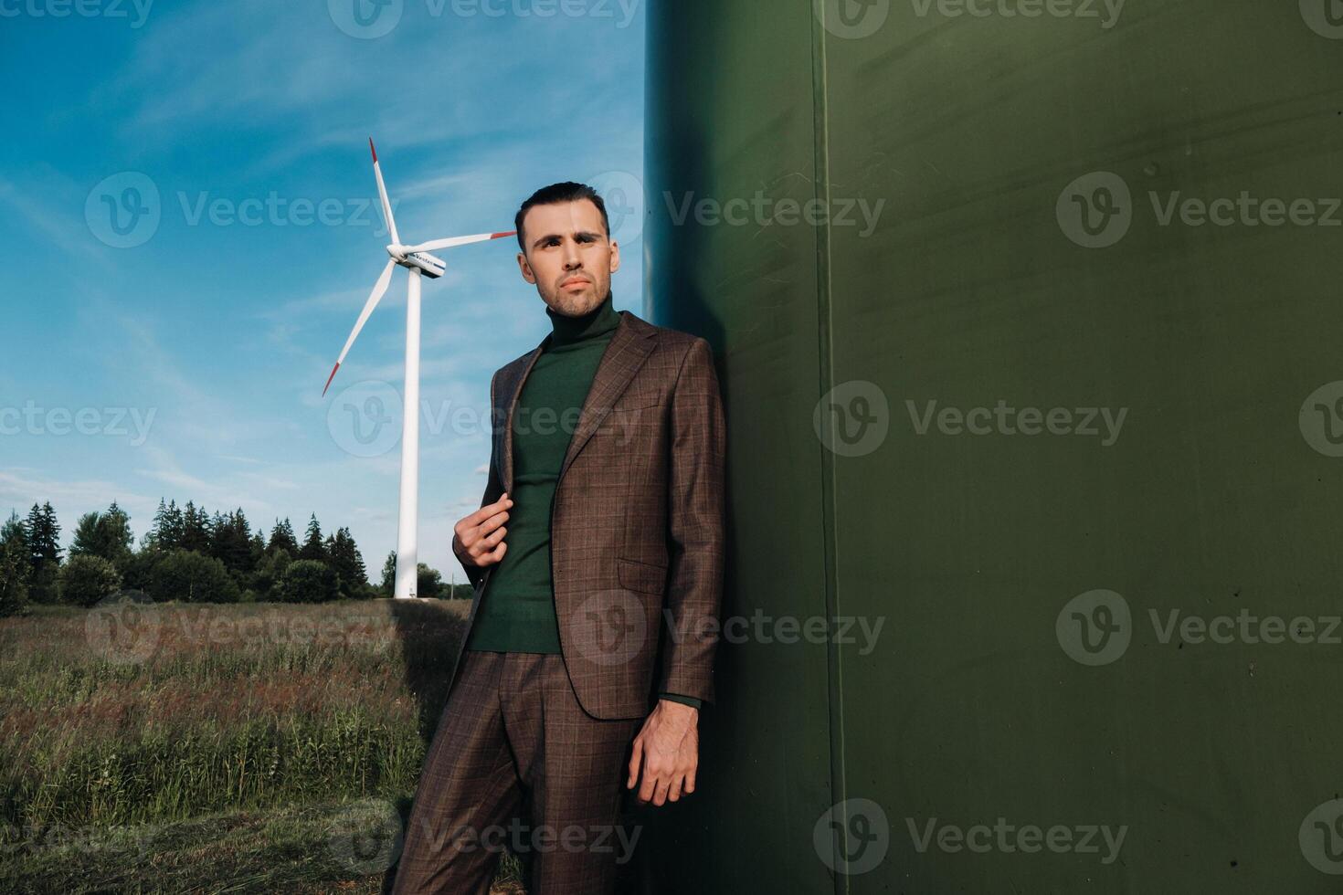 A man in a business suit with a green Golf shirt stands next to a windmill against the background of the field and the blue sky.Businessman near the windmills.Modern concept of the future. photo