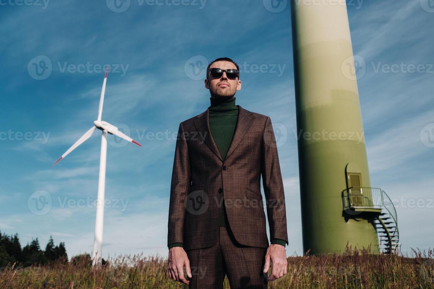 A man in a business suit with a green Golf shirt stands next to a windmill against the background of the field and the blue sky.Businessman near the windmills.Modern concept of the future. photo