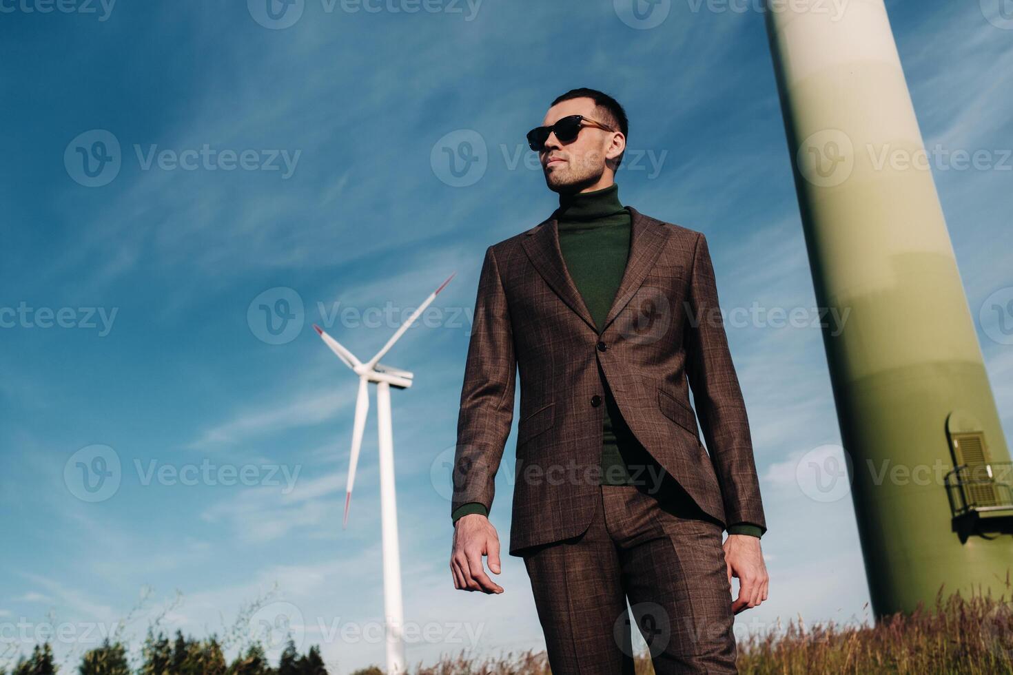 A man in a business suit with a green Golf shirt stands next to a windmill against the background of the field and the blue sky.Businessman near the windmills.Modern concept of the future. photo