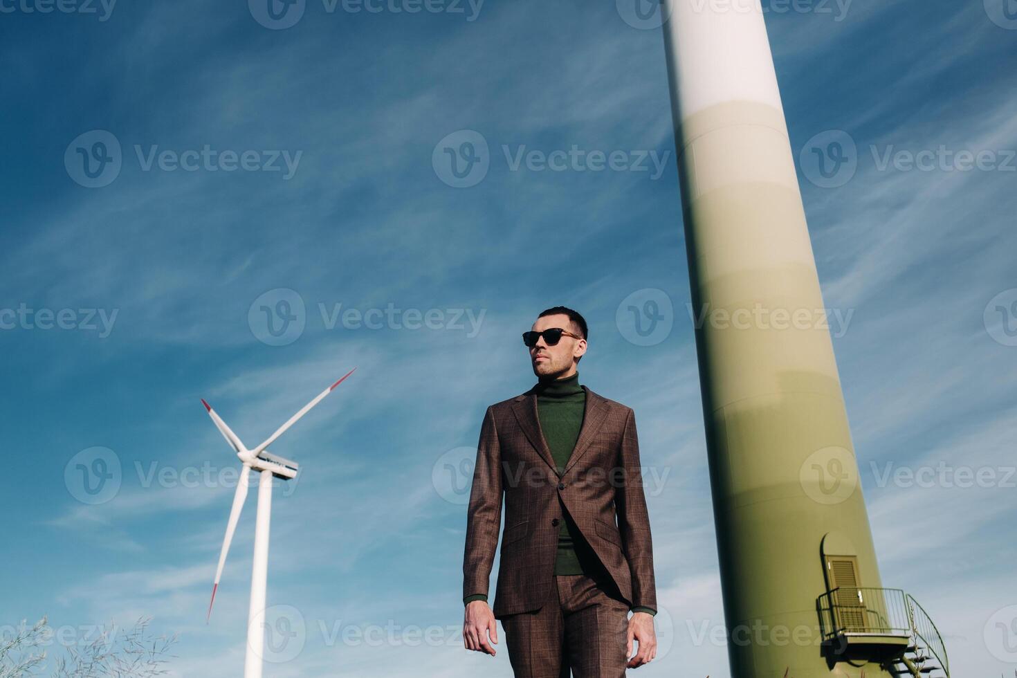 A man in a business suit with a green Golf shirt stands next to a windmill against the background of the field and the blue sky.Businessman near the windmills.Modern concept of the future. photo